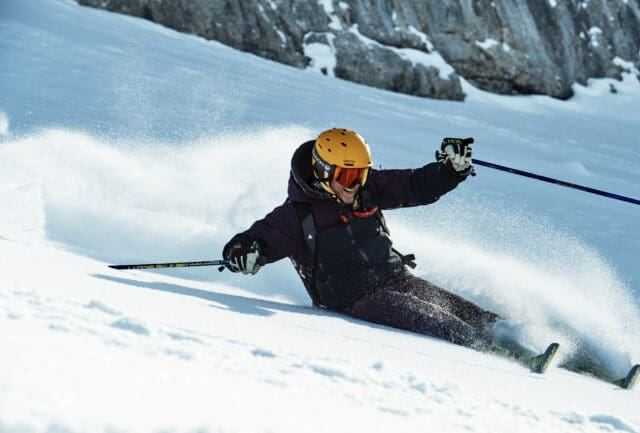 Skier in a yellow helmet leaning into a powder turn below a rock face