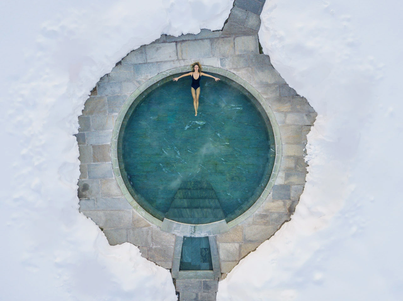 a round pool or hot tub is photographed from above, surrounded by snow, with a woman relaxing in the water