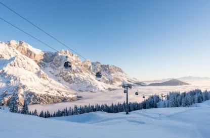 a snow dusted mountain ridge/massif behind a valley filled with clouds and a gondola travelling over snowy ski slopes