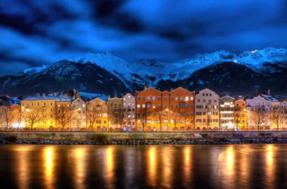 a colourful line of houses lit up, lights reflecting off the river in front, with snowy mountains as a backdrop
