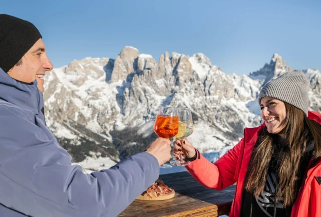 snow dusted Dolomite mountains are the backdrop to a man and woman cheersing aperitivo on the mountain top