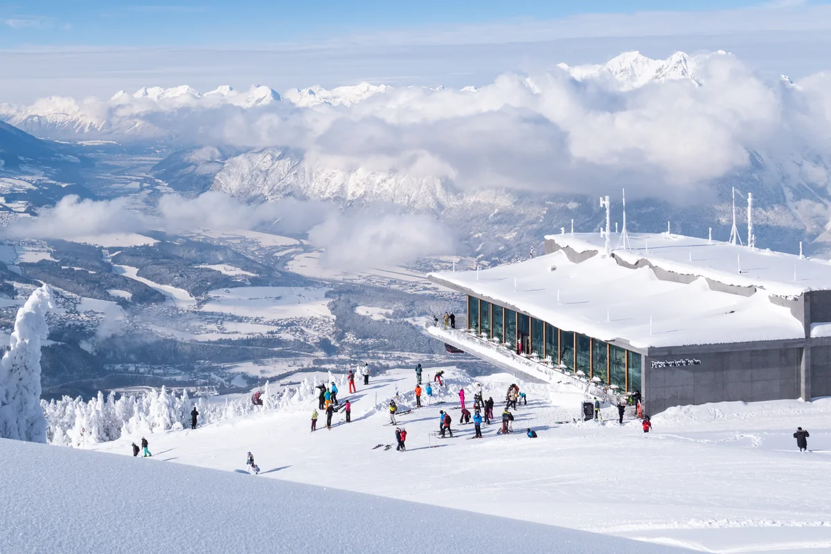 colourful skiers stand outside a mountaintop building covered in snow, the valley beautiful and snow covered below
