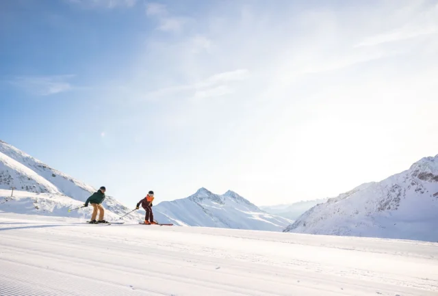 corduroy piste, crisp blue sky, two skiers skating along a high mountain flat piste