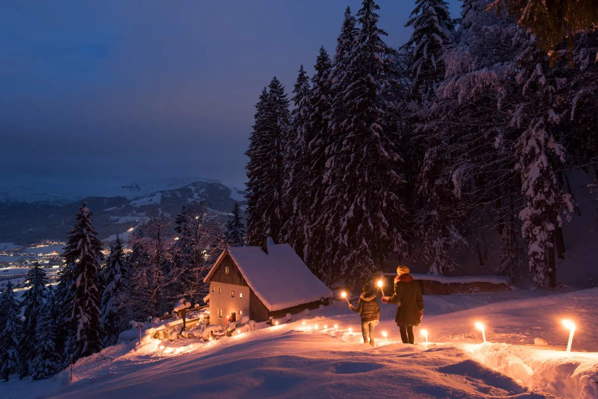 nighttime, two walkers walk down a gentle slope lit by atmospheric lights towards a snow covered cabin