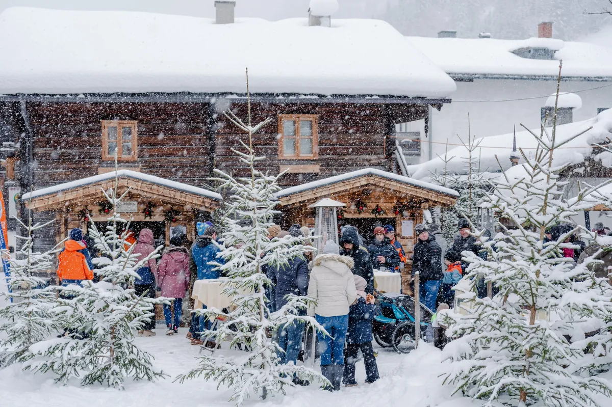 People gathered at wooden stalls in a snowy alpine village, with snow-covered roofs and fir trees creating a festive winter market scene