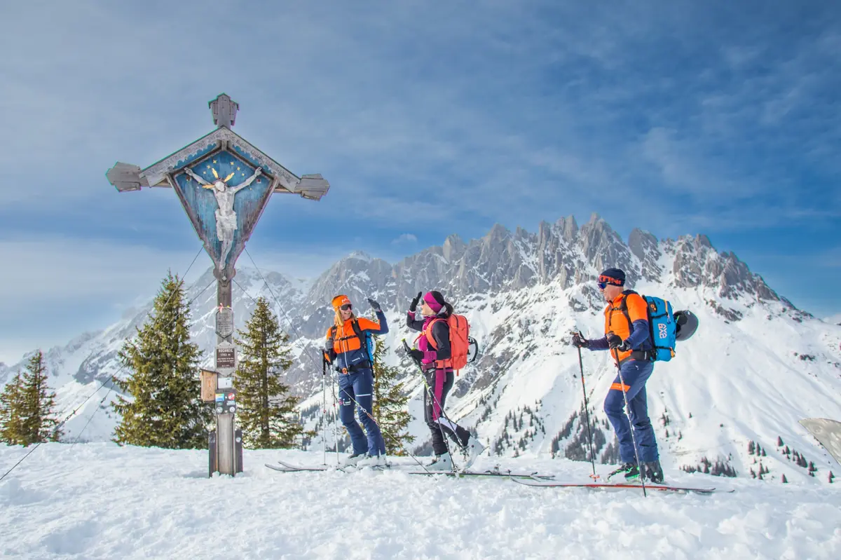 Three ski tourers in bright gear stop by a summit cross on a snowy peak in Hochkönig, with jagged alpine mountains in the background