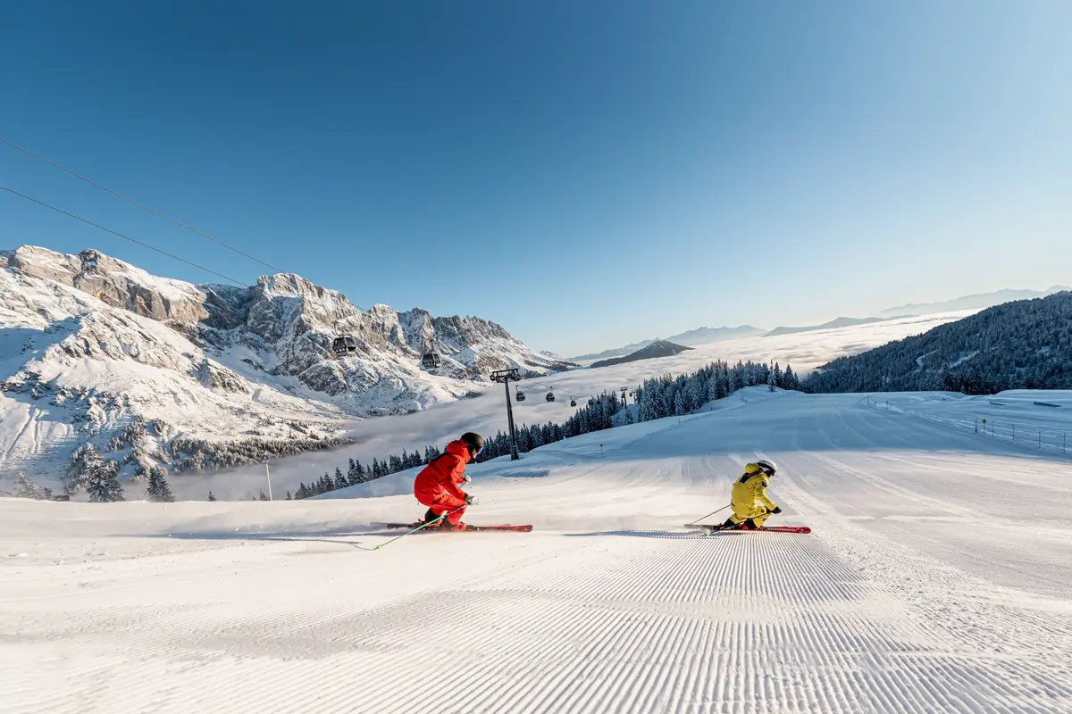 Two skiers on a pristine groomed slope with gondolas visible and dramatic mountain backdrop in Hochkönig, Austria