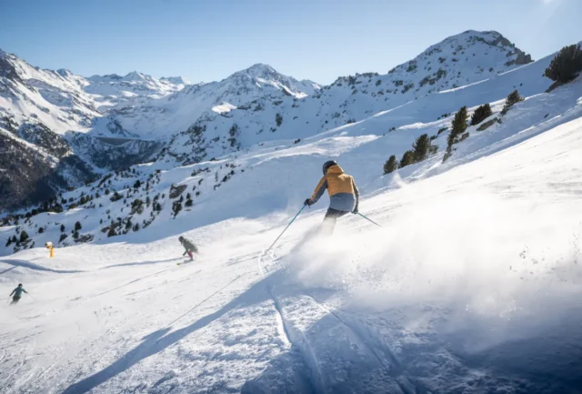 three skiers race down the piste covered in an inch of fresh snow, skis biting into the slope, the sun shining above