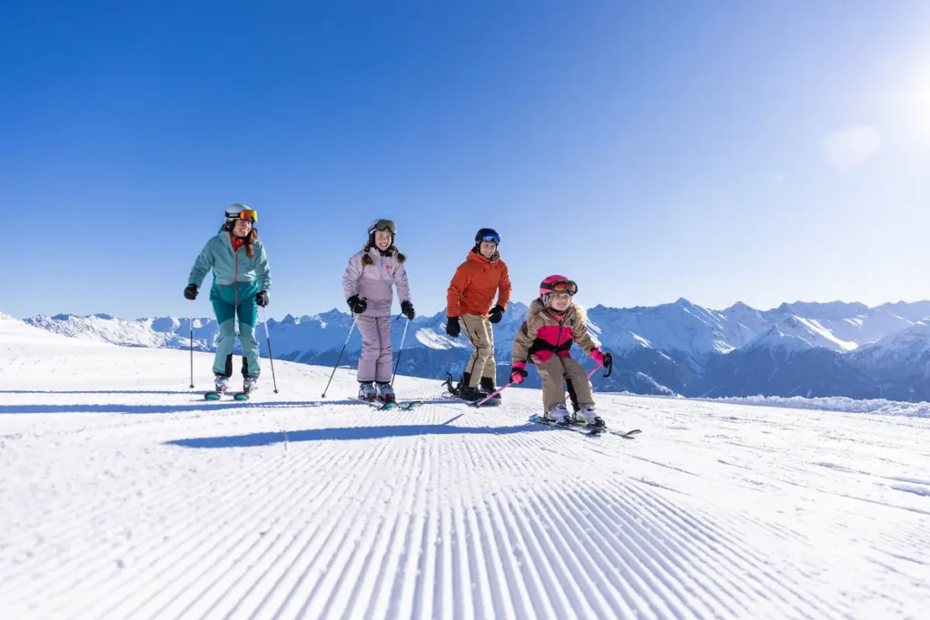 A family skiing together on a sunny groomed slope in Serfaus-Fiss-Ladis, surrounded by snow-covered Alpine peaks under a clear blue sky.