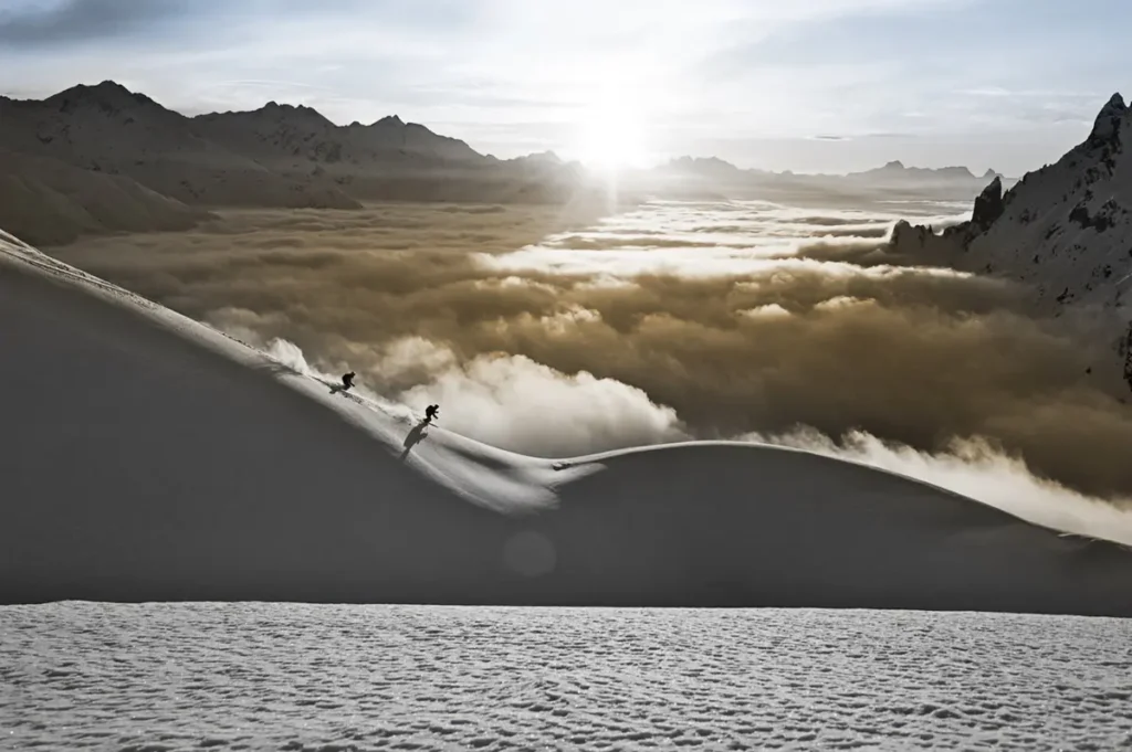 Two skiers descend a high alpine ridge above a sea of clouds in Lech Zรผrs am Arlberg at sunrise, framed by jagged peaks.