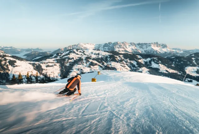 A skier in red and black races down a perfectly groomed slope in Ischgl, snow spraying in bright sunlight against blue skies