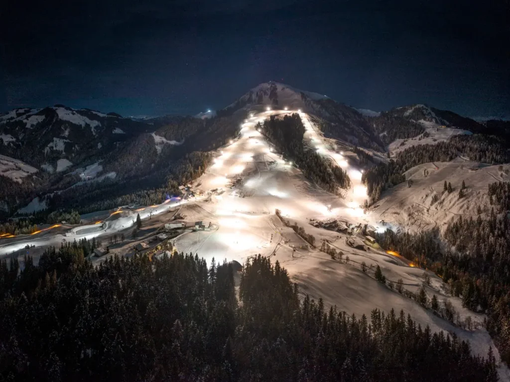 An aerial view of Sรถllโs floodlit ski slopes at night in the SkiWelt Wilder Kaiser region, glowing warmly against dark forests.