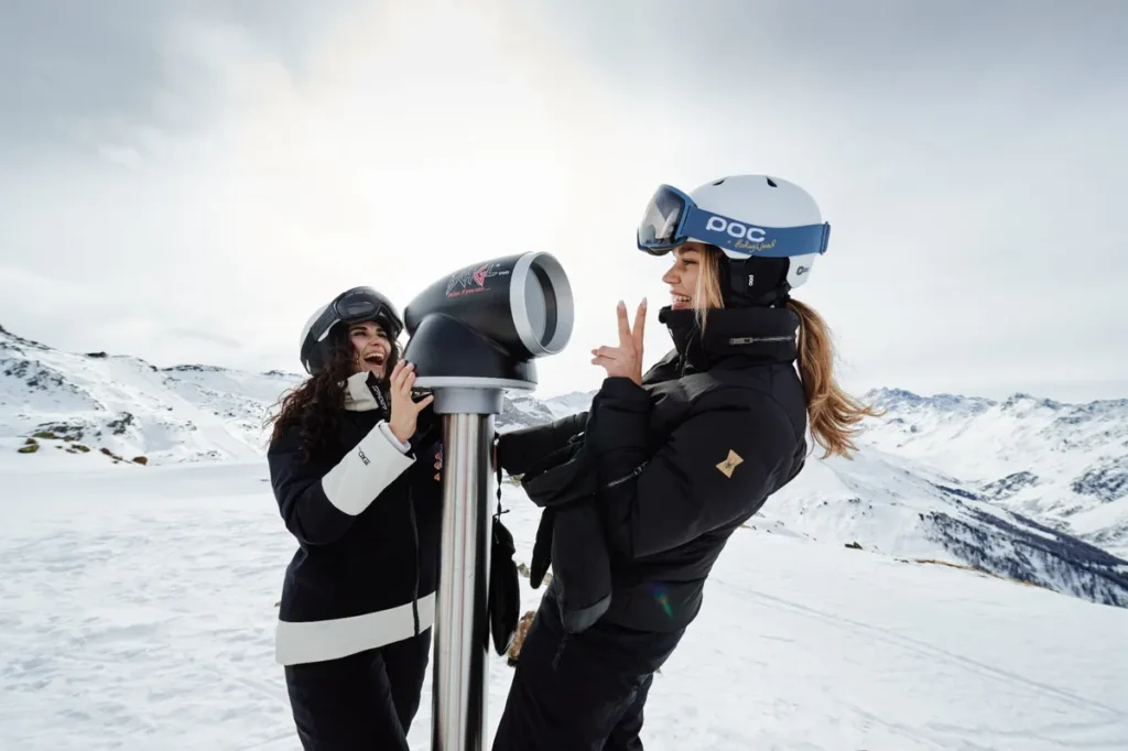 two women in ski gear, play around in front of a landscape telescope in a snowy setting