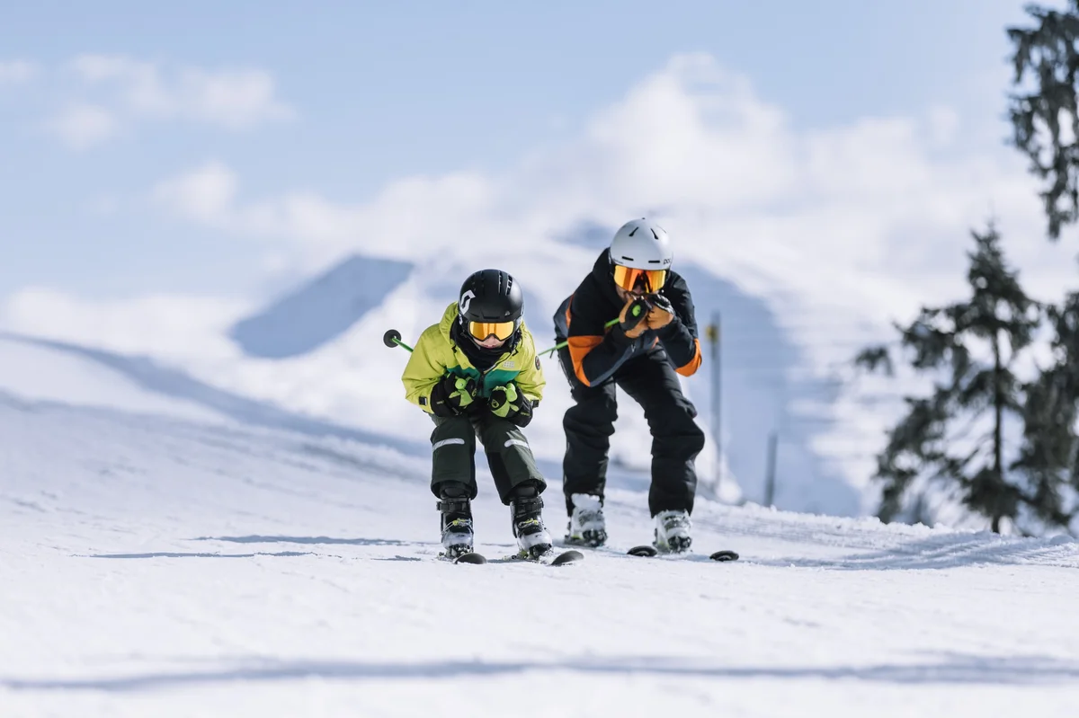 One young skier and their dad, in a racing tuck, racing down a groomed slope on a sunny winter day in Saalbach, wearing helmets and ski gear, with snowy mountains in the background.