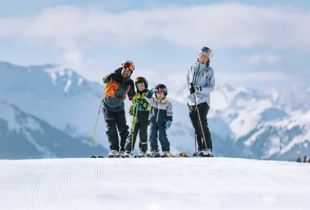 Family skiing together on a groomed slope in Saalbach, with panoramic alpine peaks behind them on a clear winter day.
