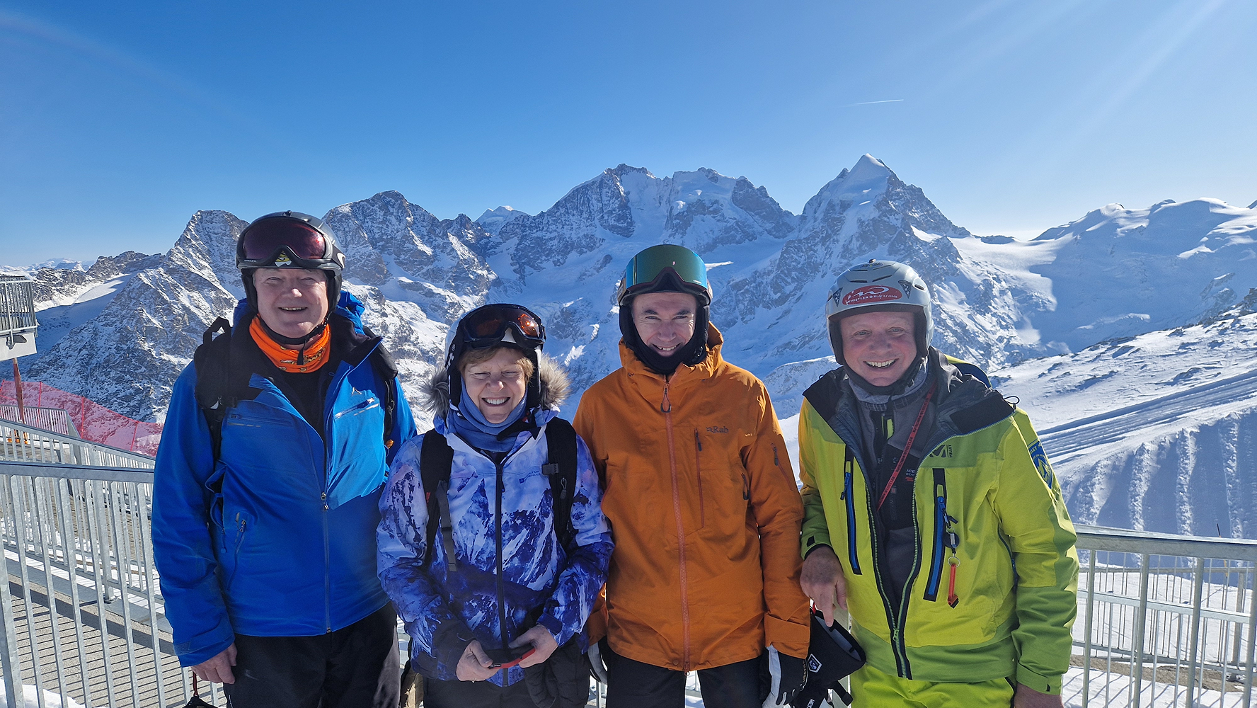 Four Ski Club members smiling together on a viewing platform above snowy peaks