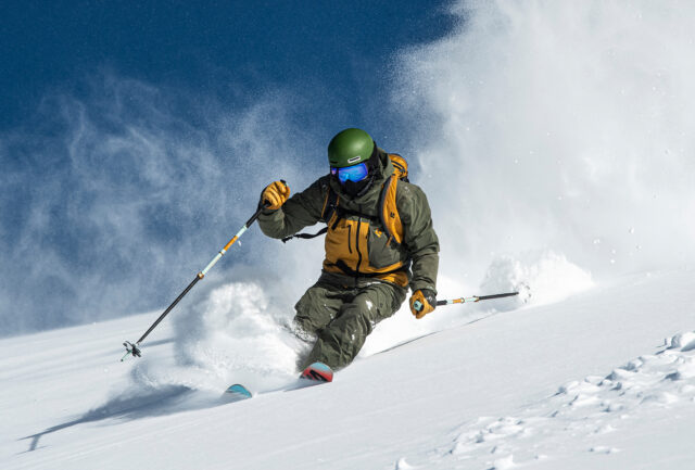 Skier in a green helmet carving through deep powder under a blue sky