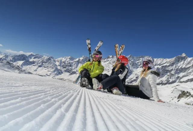 Skiers taking a break on a groomed slope with the snow-covered peaks of the Aosta Valley behind them.