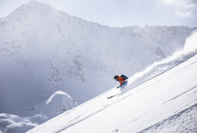 Skier making a powder turn on a steep slope below snowy mountains