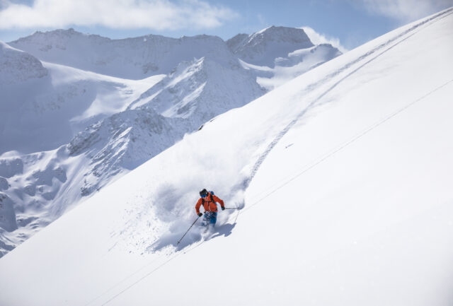 Skier in an orange jacket carving fresh powder high in the mountains