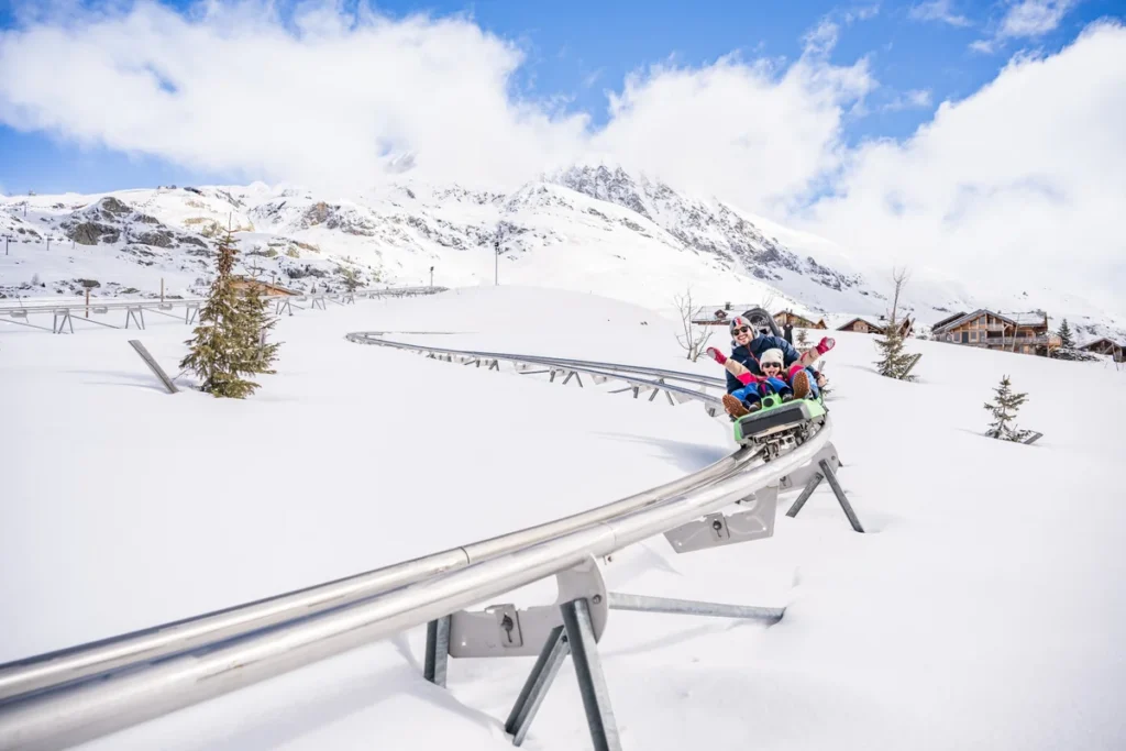 Family riding a mountain coaster through snowy Alpe d’Huez, with the snow-covered Alps in the background.